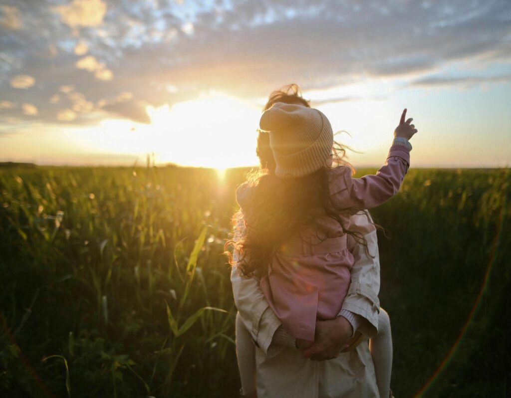 A woman holding a child stand together in a field, silhouetted against a vibrant sunset sky.